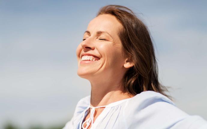 Femme brune avec haut blanc qui sourit yeux fermés avec ciel bleu en arrière-plan