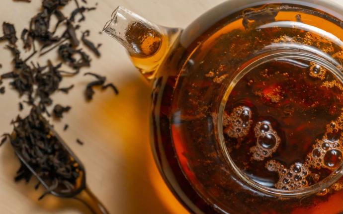 Théière transparente vue de haut avec de la tisane orangée et cuillère à côté avec tisane et feuilles séchées à côté