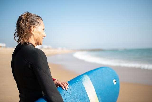 Femme chhveux courts face à la mer qui tient une planche de surf bleue