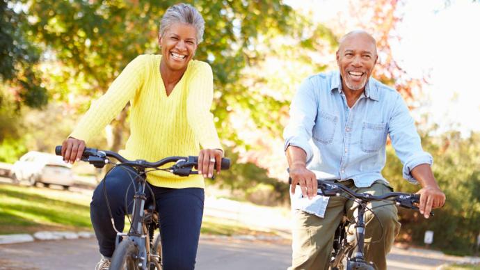 Femme senior noire avec un pull jaune et homme senior noir avec chemise en jean bleu clair qui rient sur des vélos