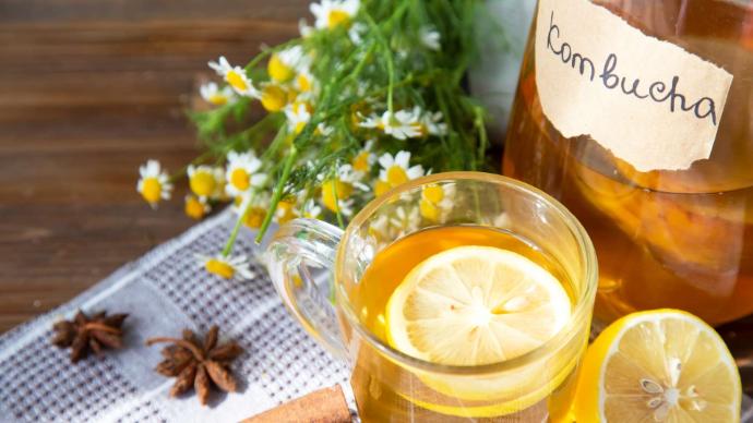 Bouteille avec liquide orange et étiquette Kombucha et un verre de kombucha avec rondelle de citron et des marguerites et étoile de cannelle à côté