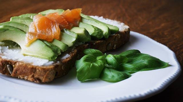 Tartine tranches d'avocats et saumon fumé avec des feuilles de basilic à côté sur une assiette blanche