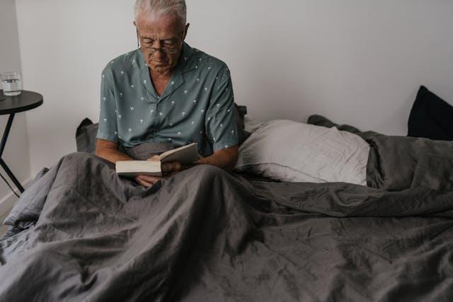 Home senior cheveux blancs et lunettes en pyjama bleu qui lit un livre dans son lit avec ses jambes sous un drap gris foncé