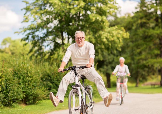 Homme senior sur un vélo les jambes écartées qui sourit avec femme senior en vélo en arrière-plan sur route bordée d'arbres