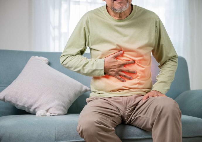 Homme avec tee-shirt manches longues vert et pantalon beige assis dans canapé bleu qui a la main sur son estomac douloureux