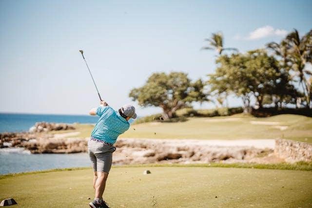 Homme de dos avec chemise bleu et short beige qui joue au golf avec mer en arrière-plan