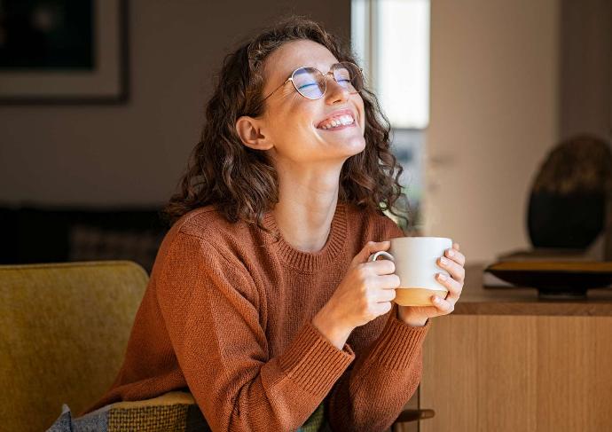Jeune femme avec lunettes et pull rouille qui sourit tête vers le haut et yeux fermés dans canapé avec une tasse blanche dans les mains