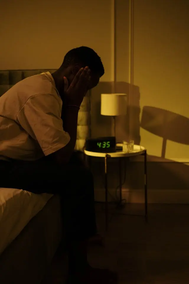 Homme noir les assis de profil au bord de son lit avec un réveil indiquant 4h35