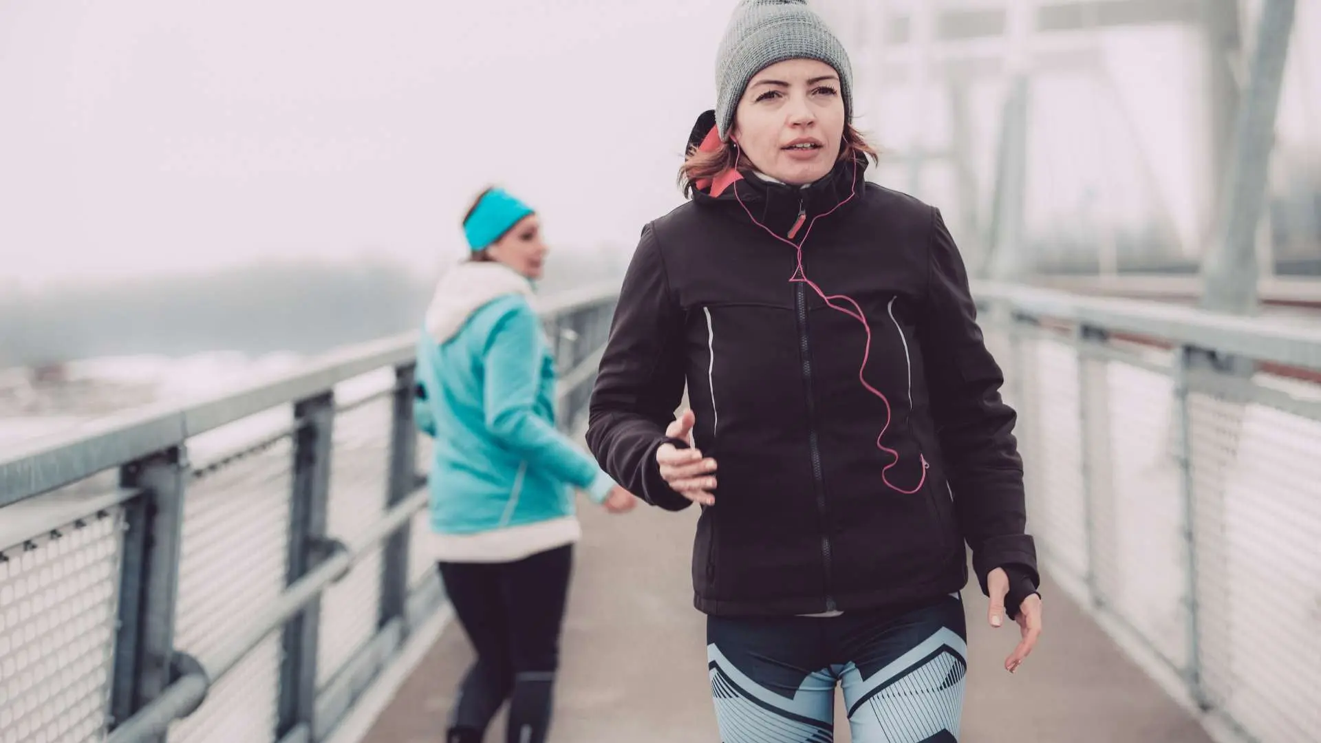 Femme en tenue de sport et bonnet qui marche sur un pont métallique avec une autre femme de trois quart dos derrière elle en tenue de sport 