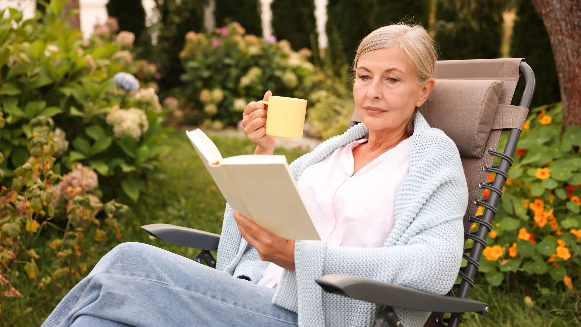 Femme senior dans une chaise longue marron qui lit un livre avec une tasse dnas l'autre main dans un jardin fleuri