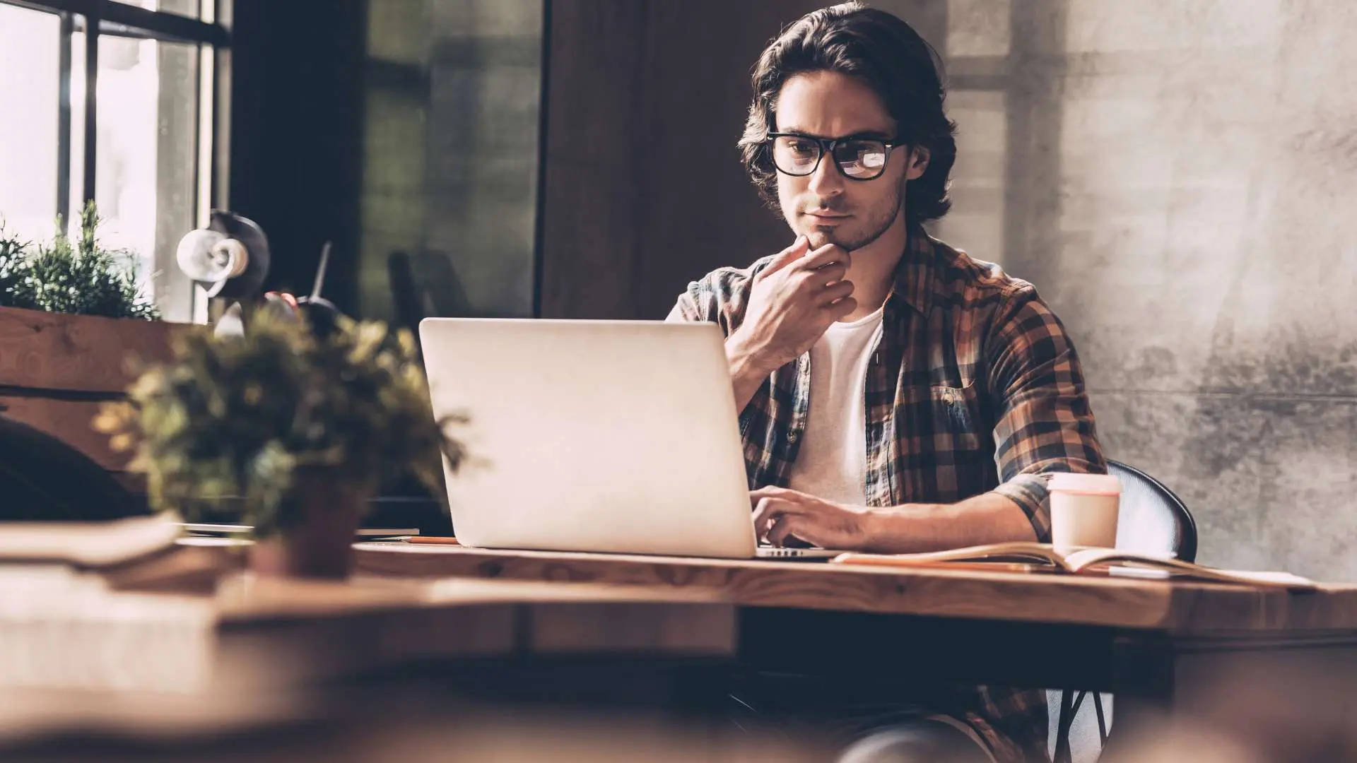 Homme à cehmise à carrezaux, tee-shirt blanc et lunettes concentré devant son ordinateur avec plante au premier-plan