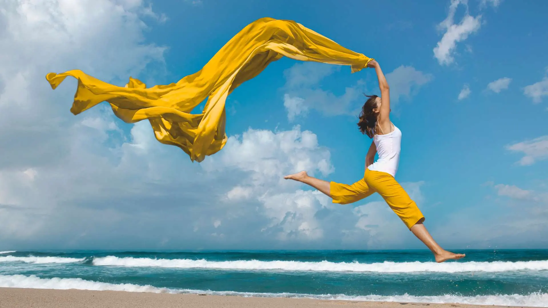 Femme qui saute avec pantalon jaune et débardeur blanc qui tient une grande étole  jaune volant derrière elle avec la plage, la mer et le ciel bleu en arrière-plan