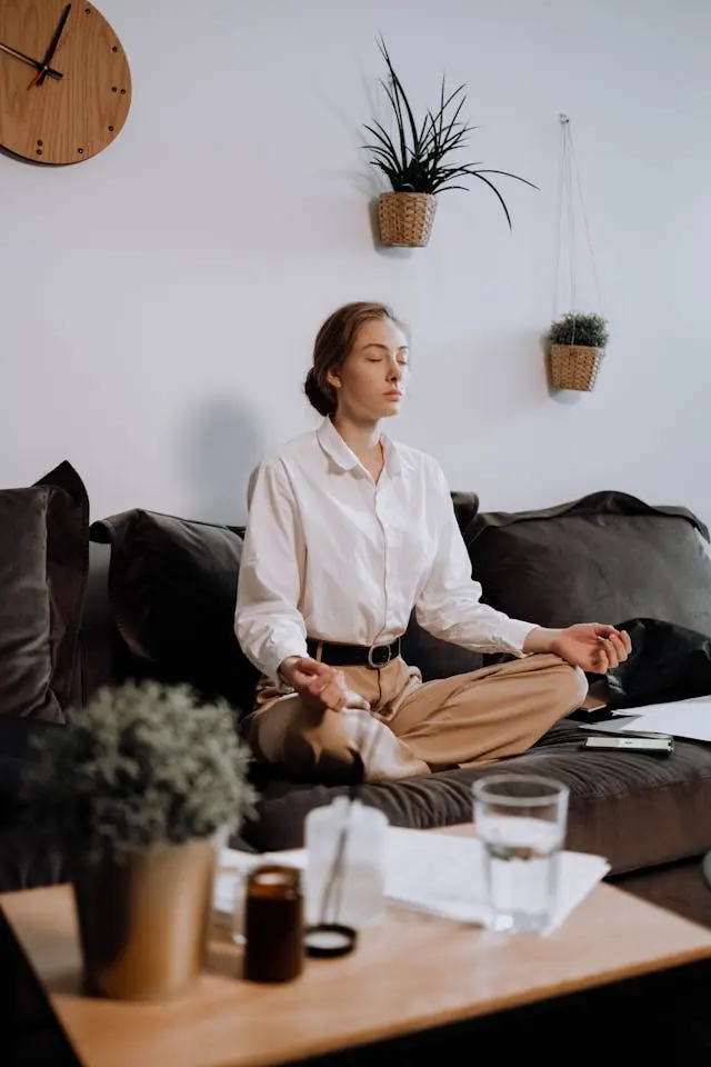 Jeune femme assise en tailleur qui médite sur canapé vert olive en velours avec table en bois, plantes et verre d'eau