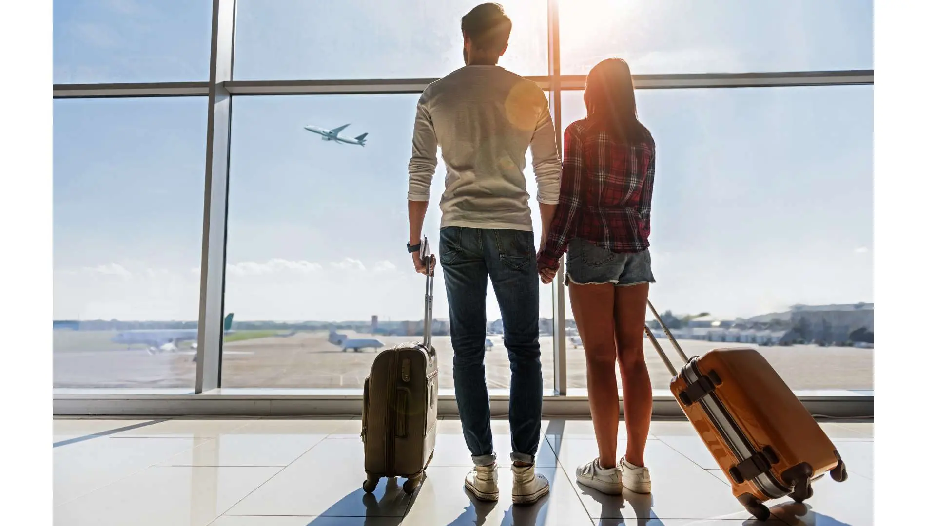 Couple de voyageurs de dos avec valise devant une baie vitrée d'aéroport qui regardent un avion décoller