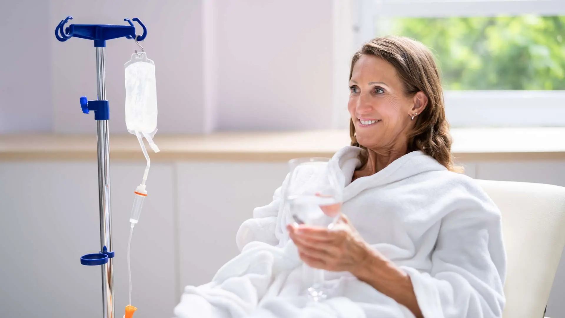 Femme en peignoir blanc verre d'eau à la main souriant pendant une séance de perfusion royale