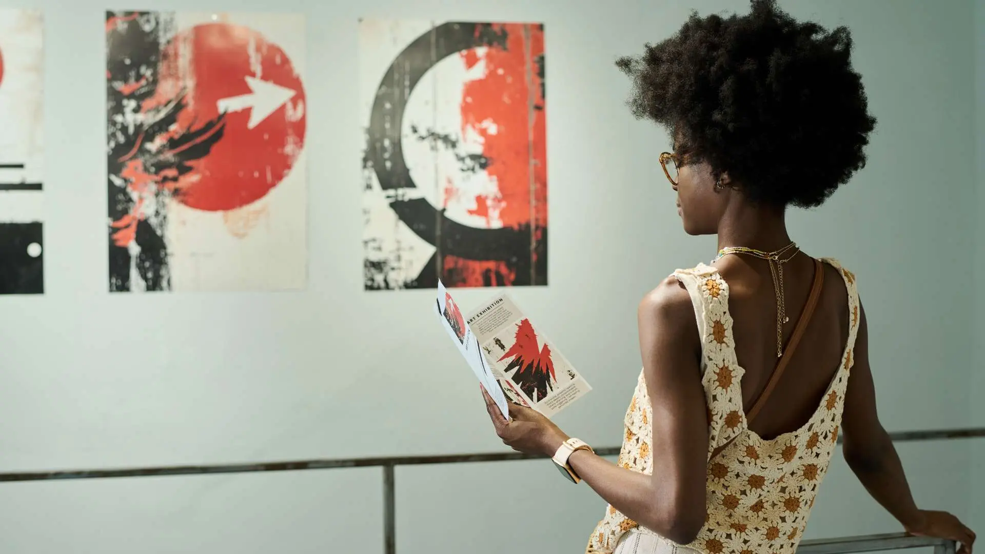 Jeune femme noire en débardeur de dos tenant un fascicule devant un tableau contemporain rouge, noir et blanc dans un musée