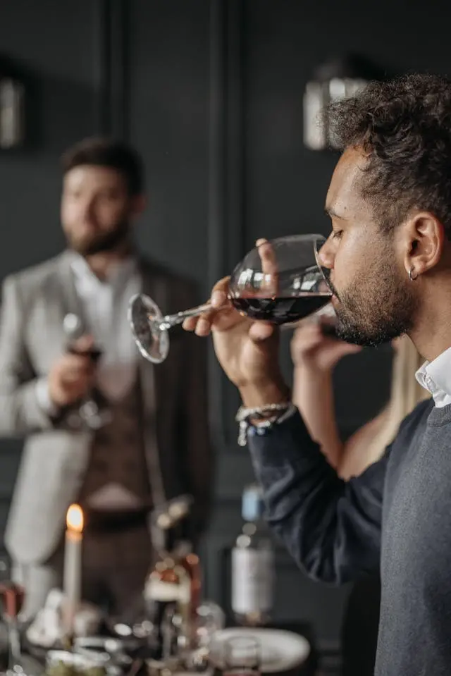 Jeune homme noire de profil qui boit du vin rouge dans un verre avec un autre homme en arrière-plan qui tient un verre de vin à la main