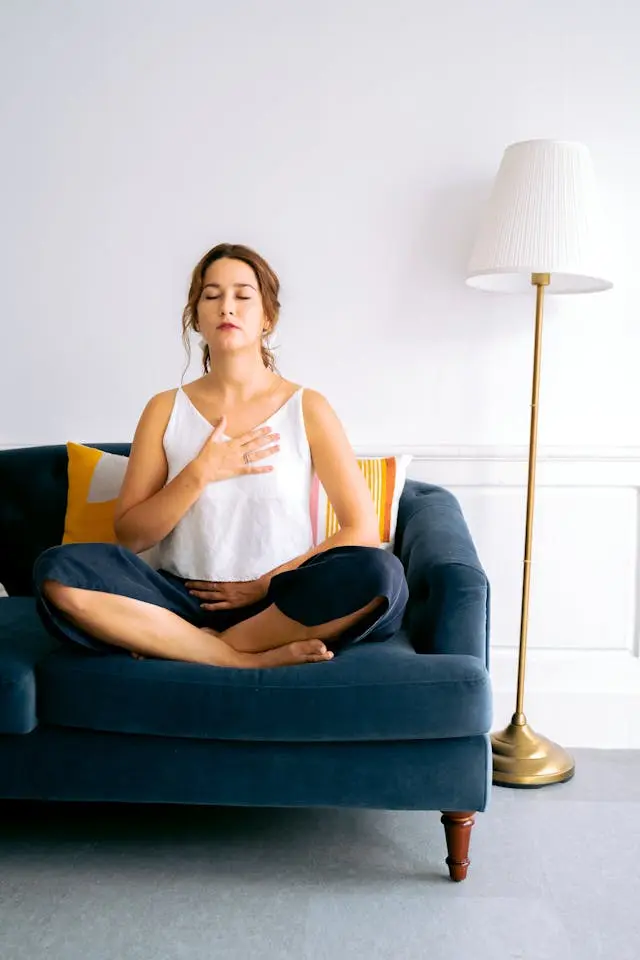 Femme assise yeux fermés en  tailleur sur un canapé bleu en velours qui respire une main  sur la cage thoracique et l'autre sur le bas du ventre