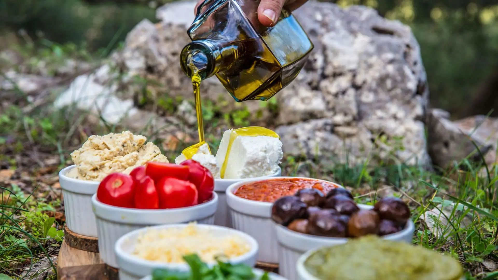 Ramequins blancs avec tomates, tapenade, olives posés dans l'herbe avec un rocher en arrière-plan et une main versant un filet d'olive sur du fromage