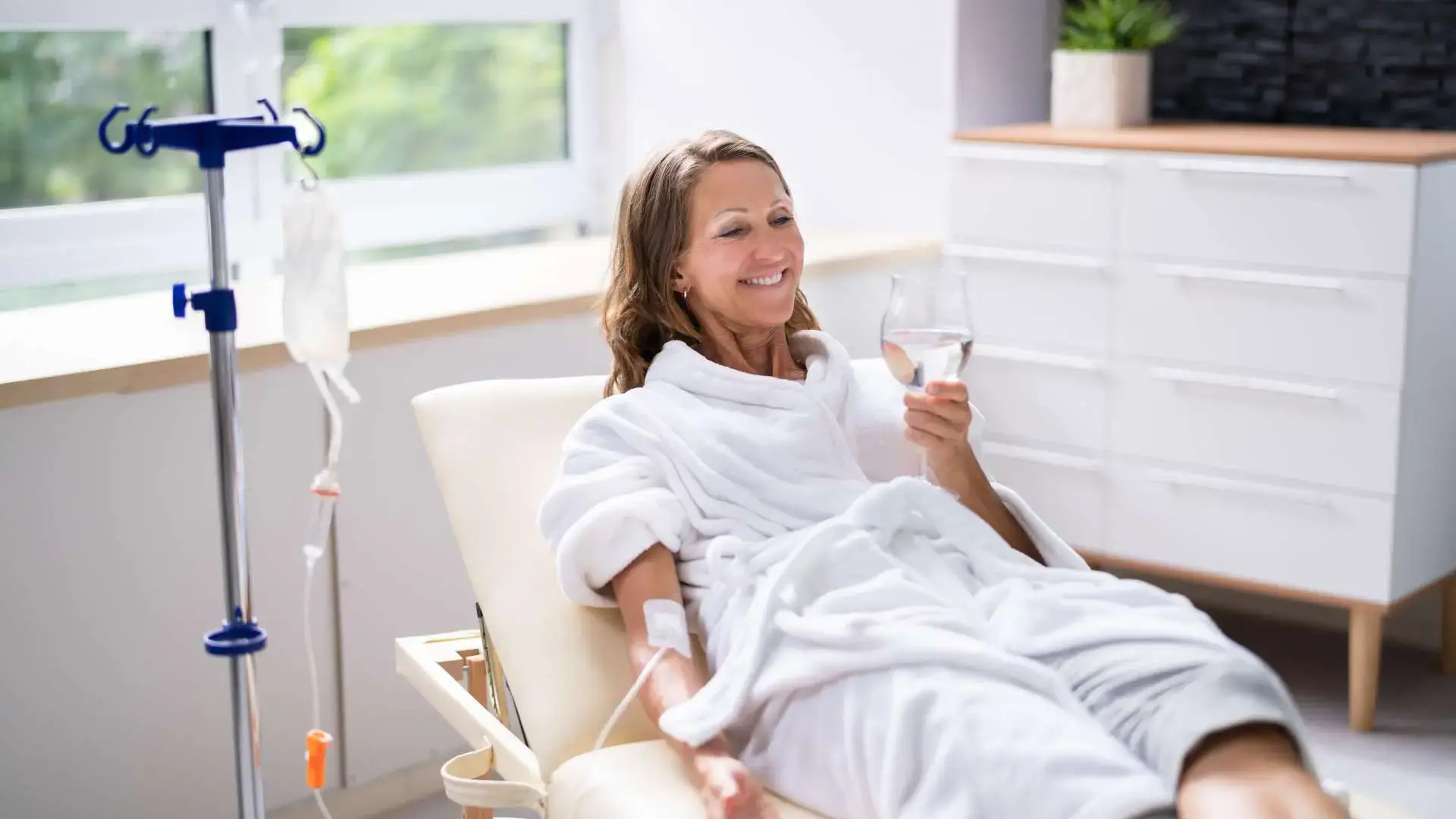 Femme en peignoir blanc souriant avec une verre d'eau à la main pendant une séance de thérapie par perfusion