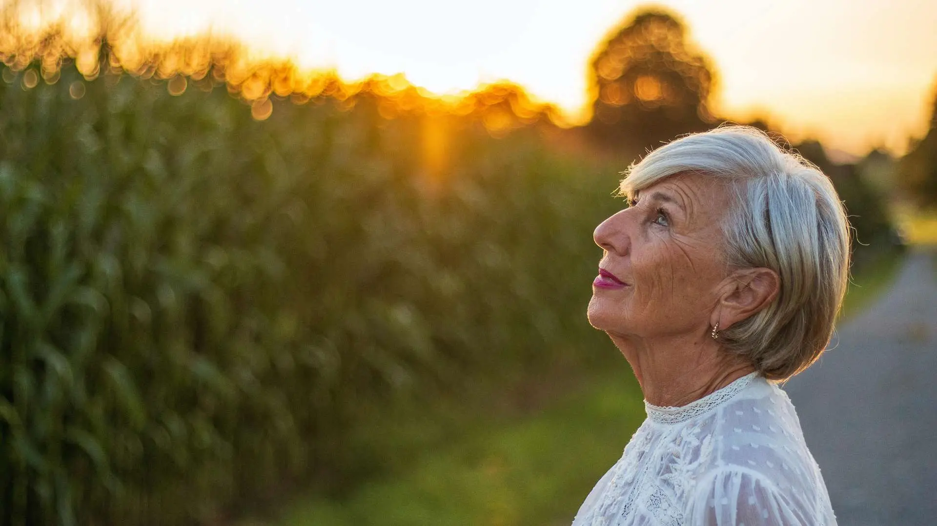 Femme senior cheveux courts blancs regardant vers le ciel en souriant devant haie verte et soleil couchant